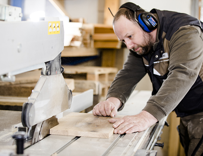 Tischler bei der präzisen Holzbearbeitung an einer Säge in der Werkstatt Siebert Tischler mit Gehörschutz arbeitet konzentriert an einer Kreissäge und bearbeitet ein massives Holzbrett